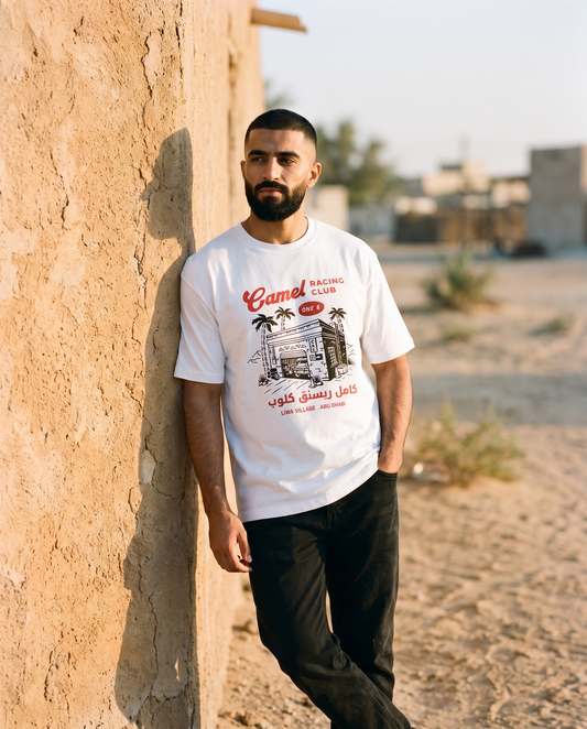 Desert Workshop Camel Racing Club tee, male model leaning on village garage wall