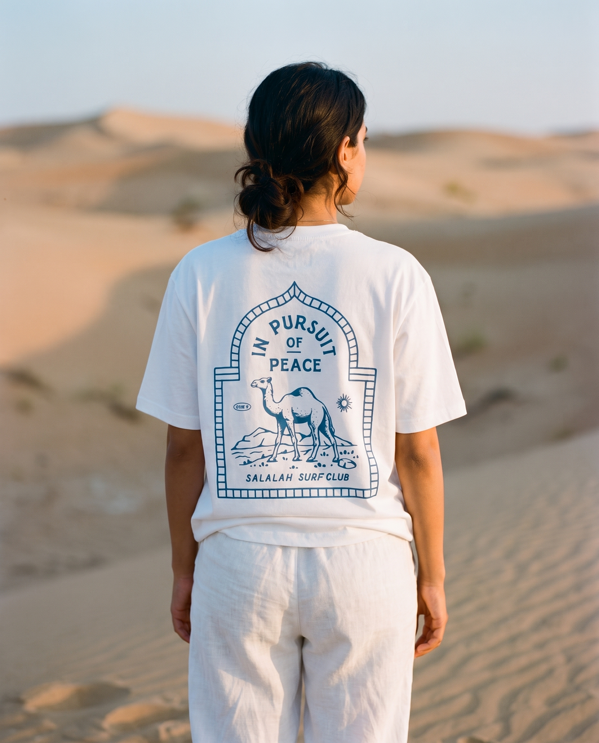 Salalah Surf Club Camel tee back print worn by female model in desert dunes