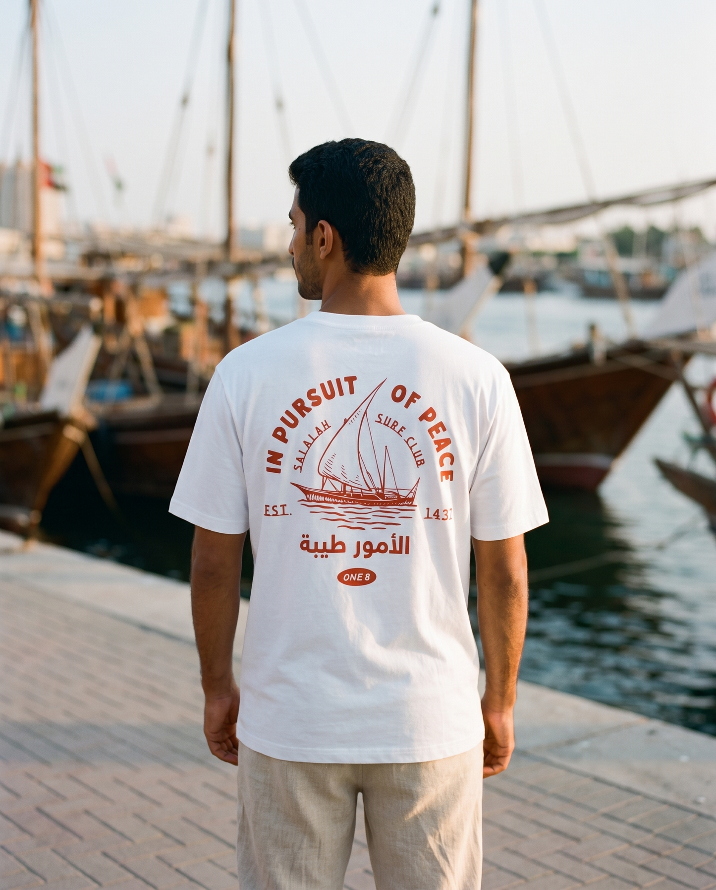 Salalah Surf Club Dhow tee back print worn by male model at Dubai Creek with dhow boats