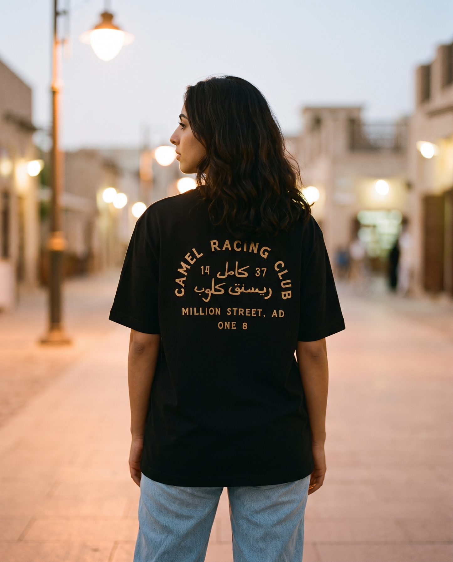 Million Street Tee Camel Racing Club gold back print worn by female model on a lamplit heritage street at dusk