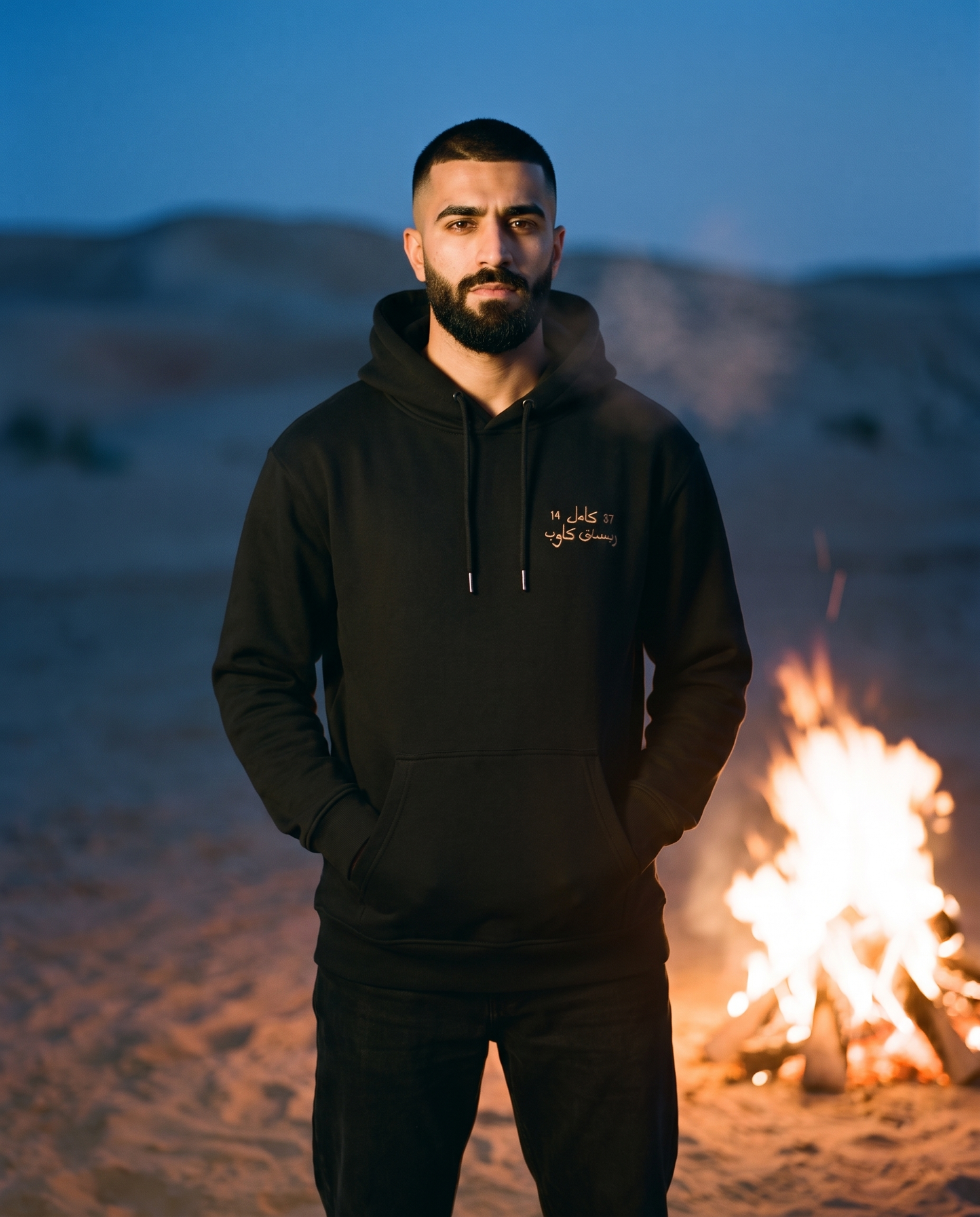 Camel Racing Club Arabic hoodie in black worn by male model beside a desert campfire at dusk