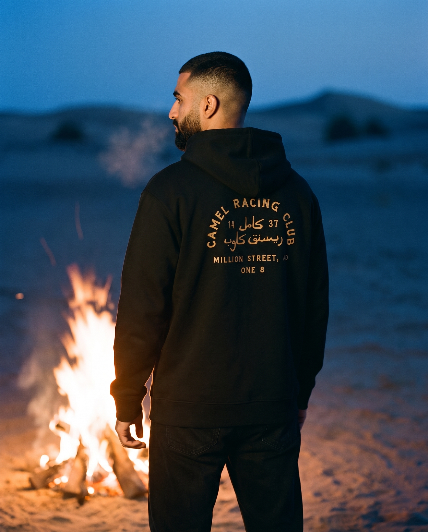 Camel Racing Club Arabic hoodie gold back print worn by male model at a desert campfire