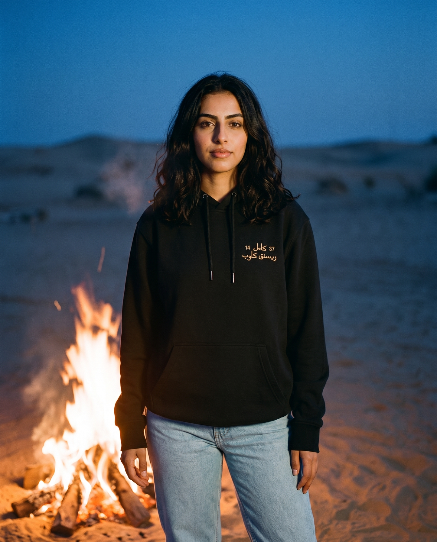 Camel Racing Club Arabic hoodie in black worn by female model beside a desert campfire at dusk