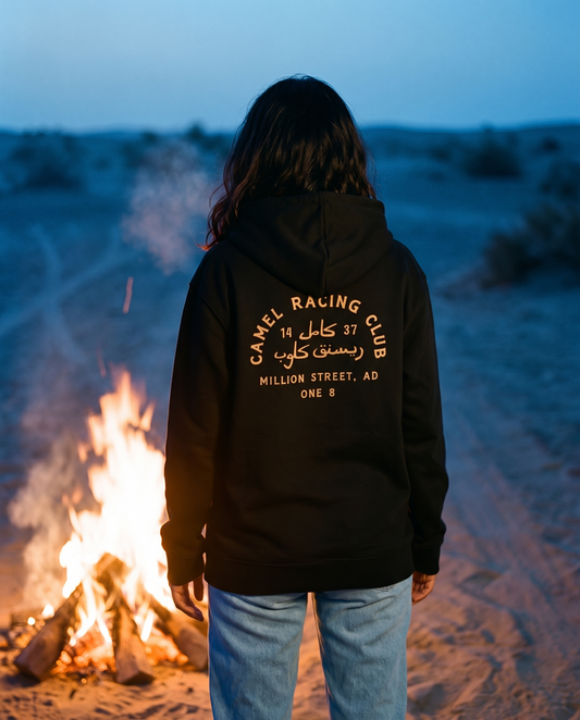 Camel Racing Club Arabic hoodie gold back print worn by female model at a desert campfire