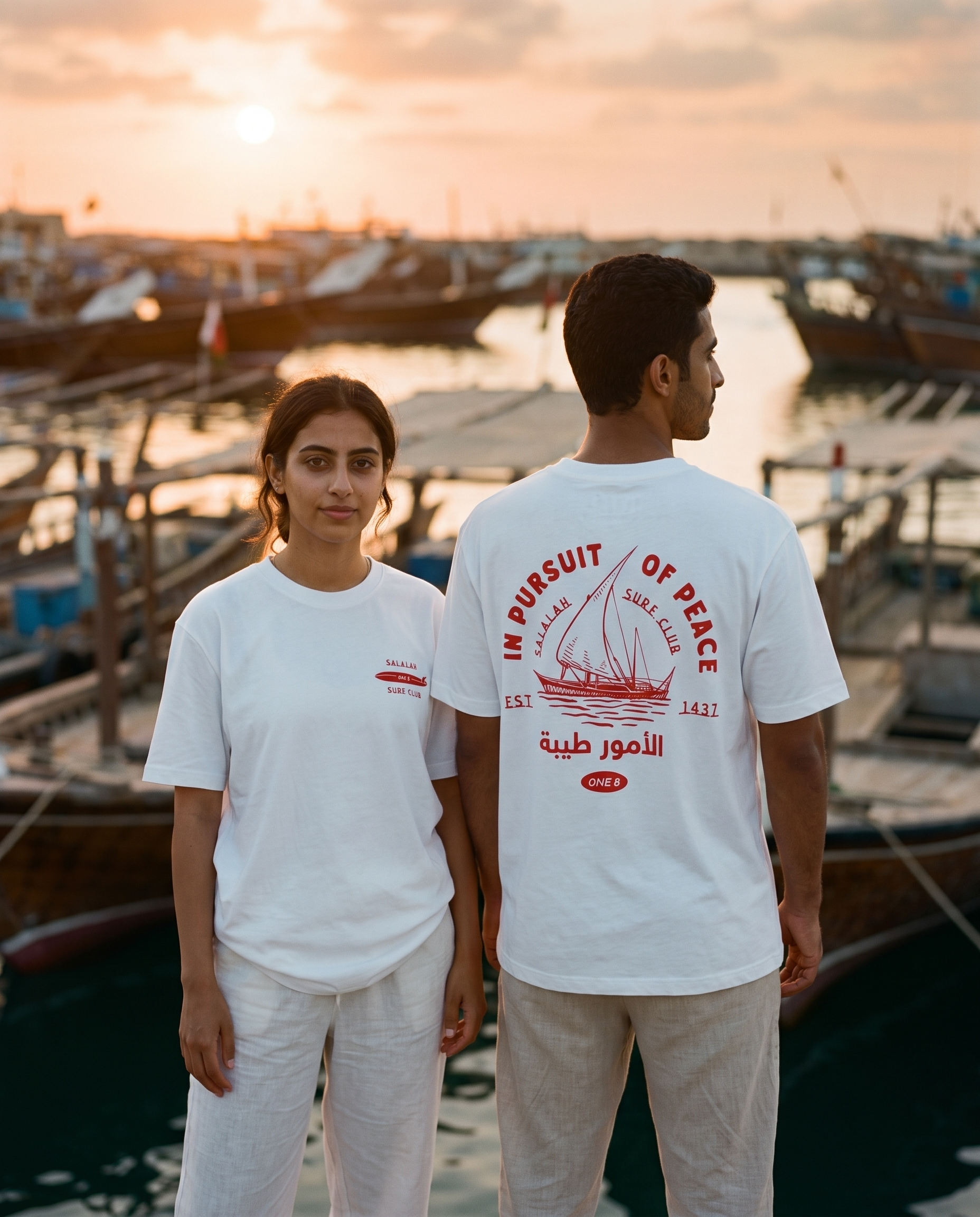 Dhow tee worn by male and female models at a dhow harbour at sunset, front and back view