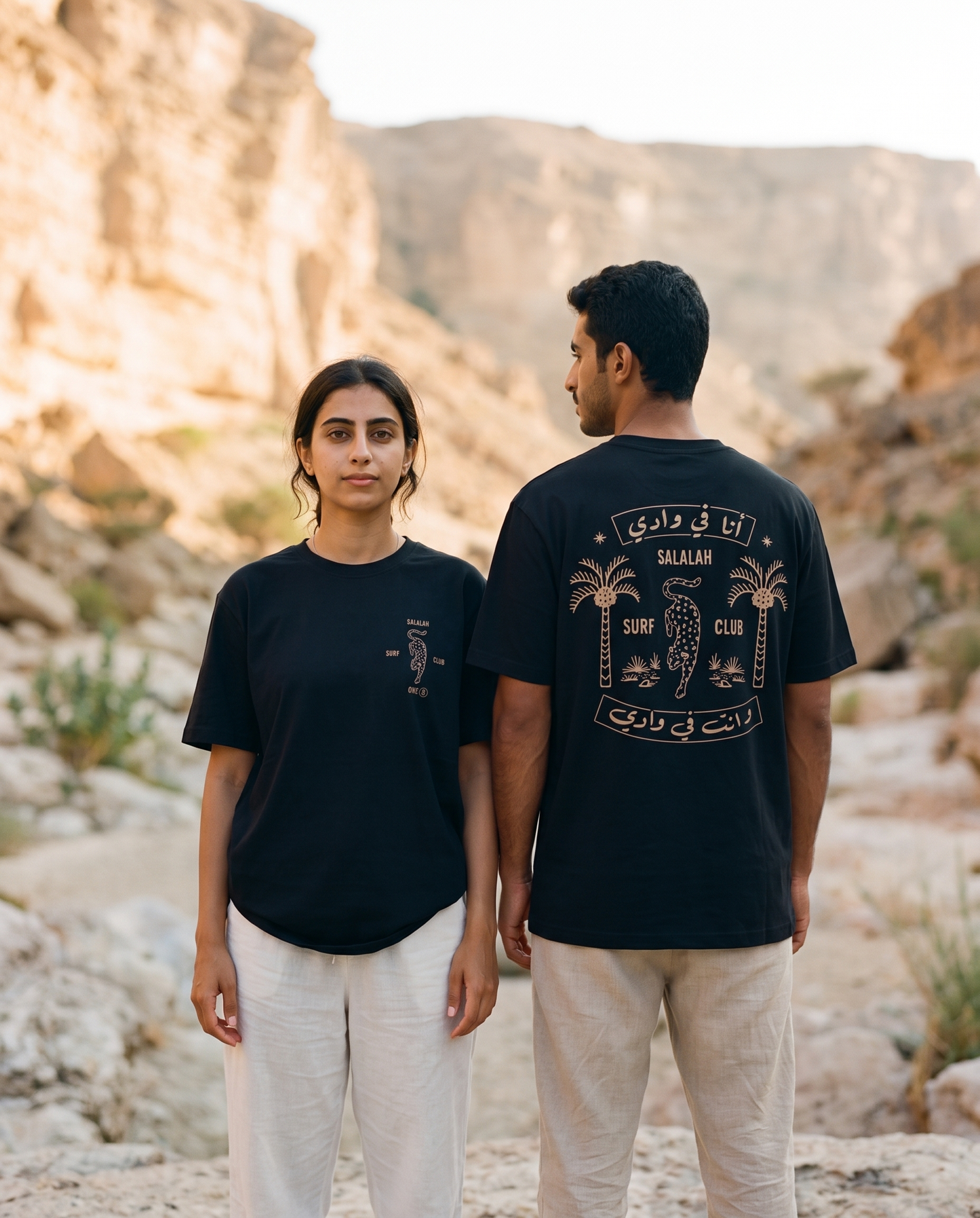 Leopard tee in black worn by male and female models in a rocky wadi, front and back view
