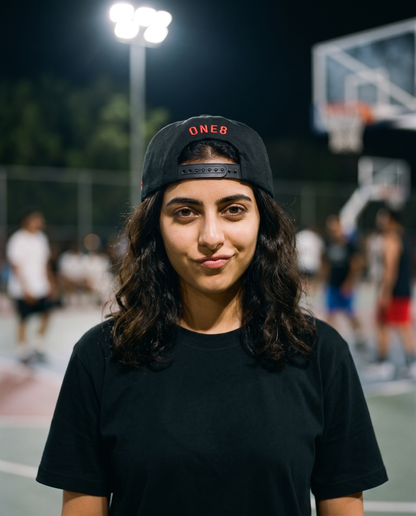 Ma'a Nafsak snapback worn backwards showing ONE8 back, female model on floodlit court